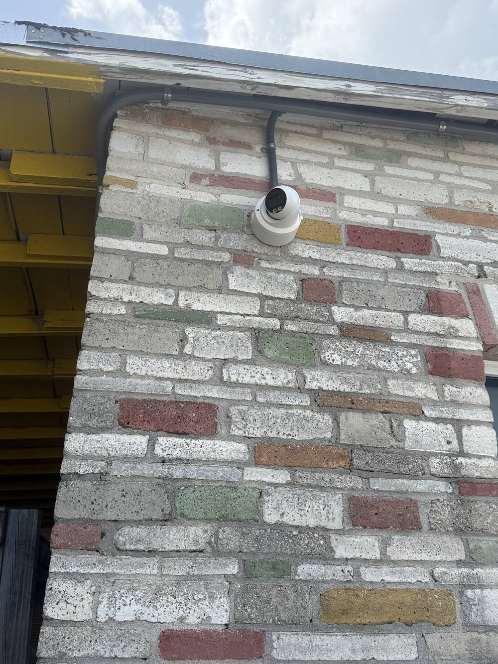 A dome security camera mounted under the roofline of the building, wired in conduit, overlooking the yard.