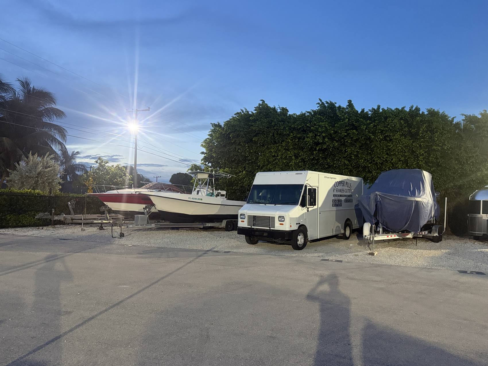 A box truck, a covered boat and center consoles parked under a lit pole light at nightfall.