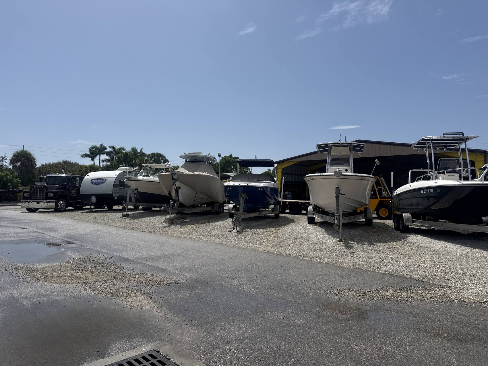 A row of center consoles and covered boats on trailers next to a Salem travel trailer and a Mack truck at the Hypoluxo Dry Dock yard.